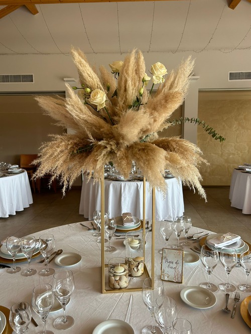 Centro de mesa alto com estrutura dourada, decorado com capim-dos-pampas, rosas brancas e folhagem. Na base, conchas dentro da estrutura complementam a decoração. A mesa tem toalha branca, louça refinada e copos de vidro, num ambiente elegante e bem iluminado.